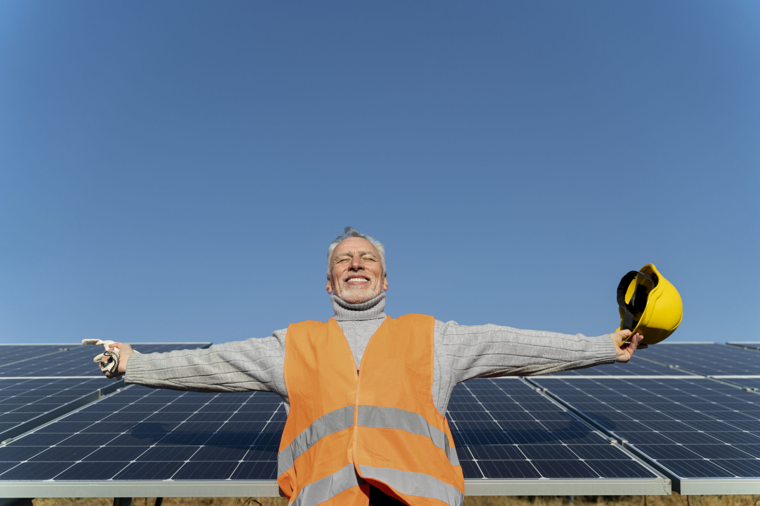 Homem grisalho, de meia-idade, vestindo um colete de segurança laranja e suéter cinza, com os braços abertos em sinal de contentamento ou sucesso, enquanto segura um capacete de segurança amarelo. Ele está em frente a painéis solares sob um céu azul claro, simbolizando a conclusão ou o sucesso de um projeto de energia renovável.