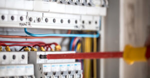 Close-up de um eletricista realizando a manutenção em um quadro de distribuição elétrica. Uma chave de fenda com isolamento vermelho é usada para ajustar um disjuntor em uma fileira organizada de componentes brancos, com fios coloridos conectados e identificados por anilhas amarelas.