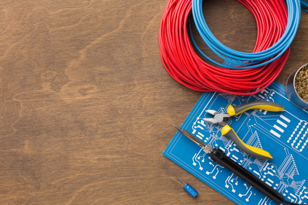 ista superior de uma mesa de madeira com materiais elétricos organizados: rolos de fios isolados nas cores vermelho e azul, um alicate de corte, um ferro de solda e uma placa de circuito impresso azul. A composição destaca ferramentas de precisão e condutores para instalações elétricas.