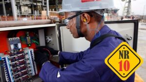 Eletricista uniformizado trabalhando em um painel elétrico externo, utilizando EPIs completos: capacete de segurança cinza, óculos de proteção transparentes, protetor auricular e luvas pretas. No canto inferior direito, há um selo amarelo de destaque com a inscrição 'NR 10'.