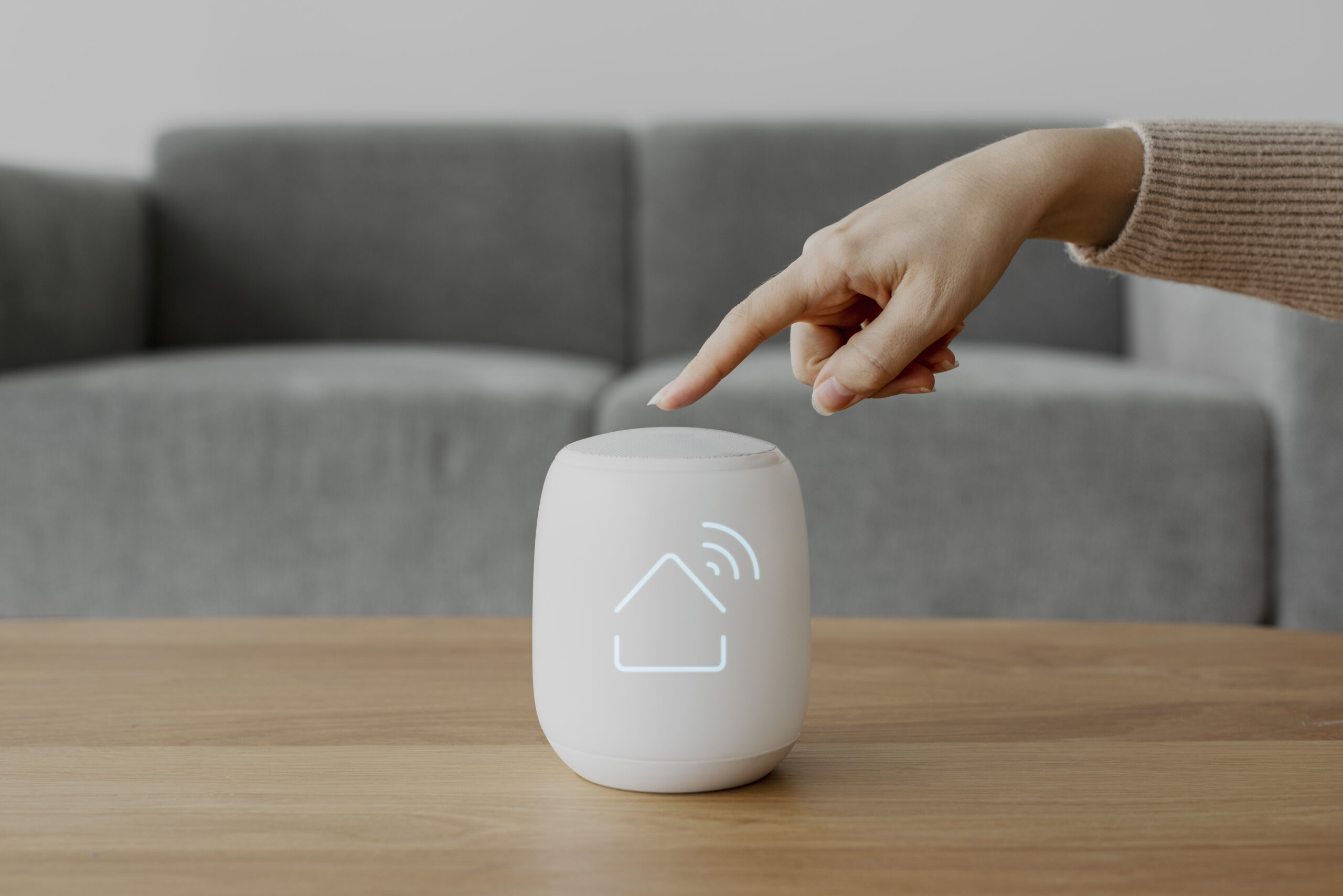 Uma mão feminina aponta para um dispositivo inteligente cilíndrico e branco sobre uma mesa de madeira. O gadget exibe um ícone brilhante de uma casa com ondas de sinal Wi-Fi, simbolizando o controle central de uma casa inteligente.