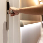 Close-up de uma mão feminina com unhas pintadas de vermelho segurando a maçaneta prateada de uma porta branca moderna. Acima da maçaneta, há um leitor de fechadura digital preta, simbolizando o uso de tecnologia smart para controle de acesso e segurança residencial.