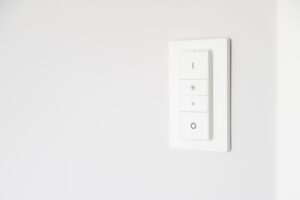 Interruptor de parede inteligente na cor branca com design minimalista, instalado em uma parede clara. O painel possui quatro botões retangulares com ícones discretos para ligar, desligar e ajustar a intensidade da luz, representando o controle intuitivo de iluminação em uma casa inteligente.