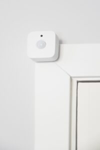 Sensor de movimento inteligente, pequeno e quadrado na cor branca, instalado no canto superior do batente de uma porta branca. O dispositivo possui uma lente circular central, exemplificando o uso de sensores discretos para automação de segurança e iluminação residencial.