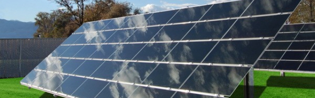 Grande instalação de painéis solares de filme fino (thin film) em solo. Os módulos possuem uma superfície lisa, escura e contínua, montados em estruturas metálicas em uma área aberta com vegetação ao fundo.