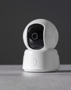 Câmera de segurança inteligente com design esférico moderno na cor branca, posicionada sobre uma superfície cinza. O dispositivo possui uma lente preta central e base giratória, exemplificando gadgets de monitoramento Wi-Fi para casas inteligentes com amplo campo de visão.