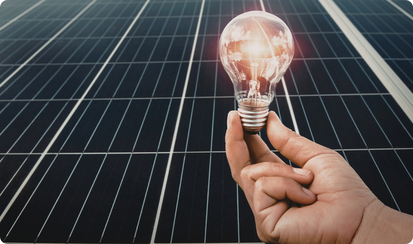Mão segurando uma lâmpada incandescente acesa com o reflexo de nuvens, contraposta a um fundo de painéis solares fotovoltaicos, simbolizando a economia de energia em Manaus e o payback da energia solar.