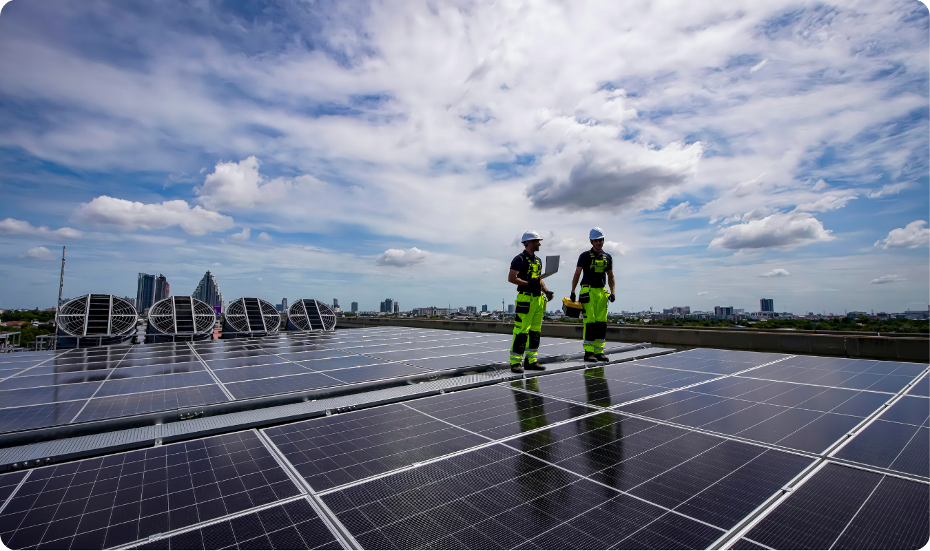 Vista panorâmica de uma instalação de energia solar em Manaus, mostrando dois técnicos com uniformes de segurança sobre uma vasta estrutura de painéis fotovoltaicos em um telhado industrial, com o skyline da cidade ao fundo sob um céu ensolarado.